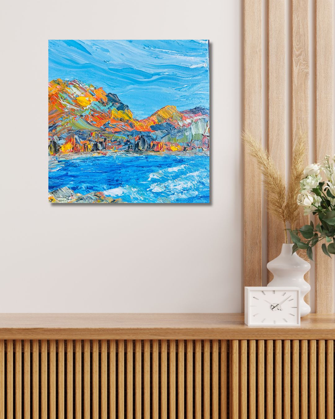 An abstract landscape painting of a blue sea and colorful mountains, by Ottawa artist Mireille Laroche, displayed on a wall in a modern interior setting with a wooden cabinet, vase, and clock.