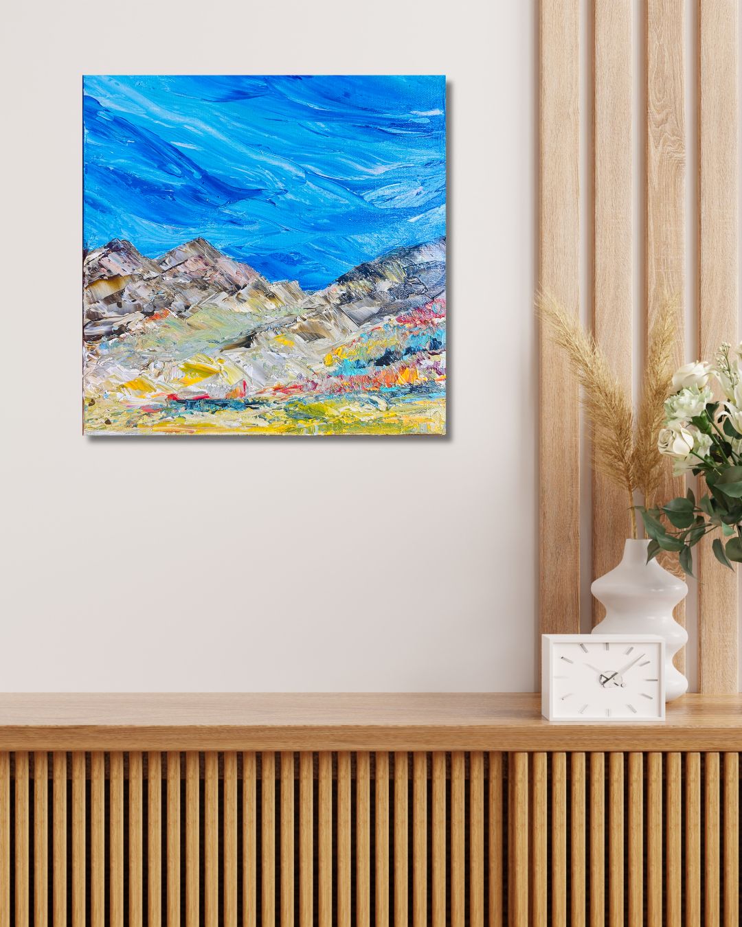 A square canvas painting of a mountain landscape with a vibrant blue sky by Ottawa artist Mireille Laroche, displayed on a light beige wall above a wooden console table with a white clock and vase of flowers.