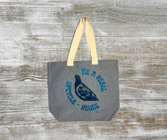 Gray tote bag with grey pigeon and blue text that says be a rebel - upcycle, reuse and a beige strap lying on a wooden surface