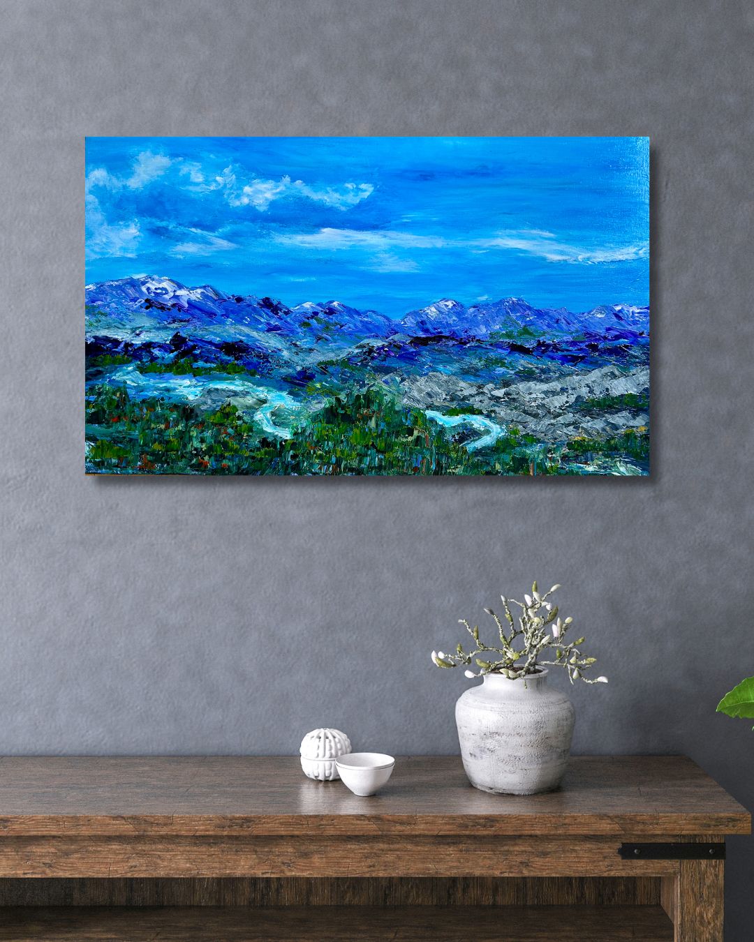 Painting of a mountain landscape  by Ottawa artist Mireille Laroche on a gray wall above a wooden table with decorative items.
