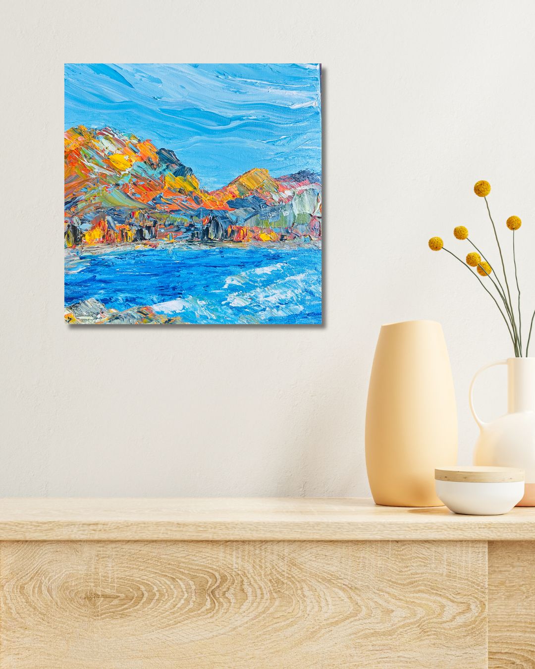 An abstract landscape painting of mountains and water in vibrant blue, orange, yellow, and green tones, by Ottawa artist Mireille Laroche, displayed on a light beige wall above a wooden console table with decorative vases.