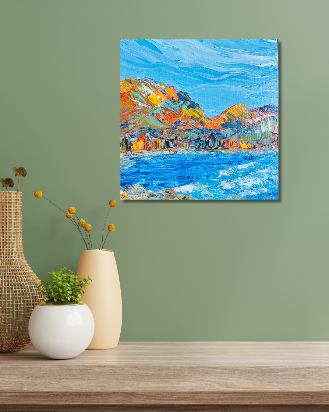 A colorful square canvas painting of a coastal landscape with blue water and orange-yellow mountains, by Ottawa artist Mireille Laroche, displayed on a sage green wall above a wooden table with decorative vases.