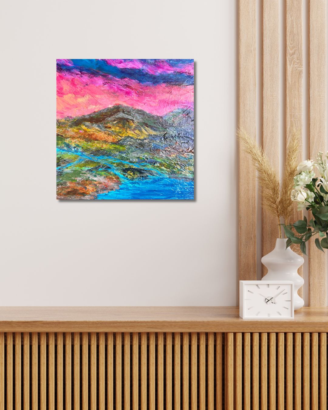 A colorful abstract landscape artwork by Ottawa artist Mireille Laroche, hanging on a light beige wall above a wooden slatted console table. The artwork features vibrant pink, purple, blue, green, and yellow brushstrokes.