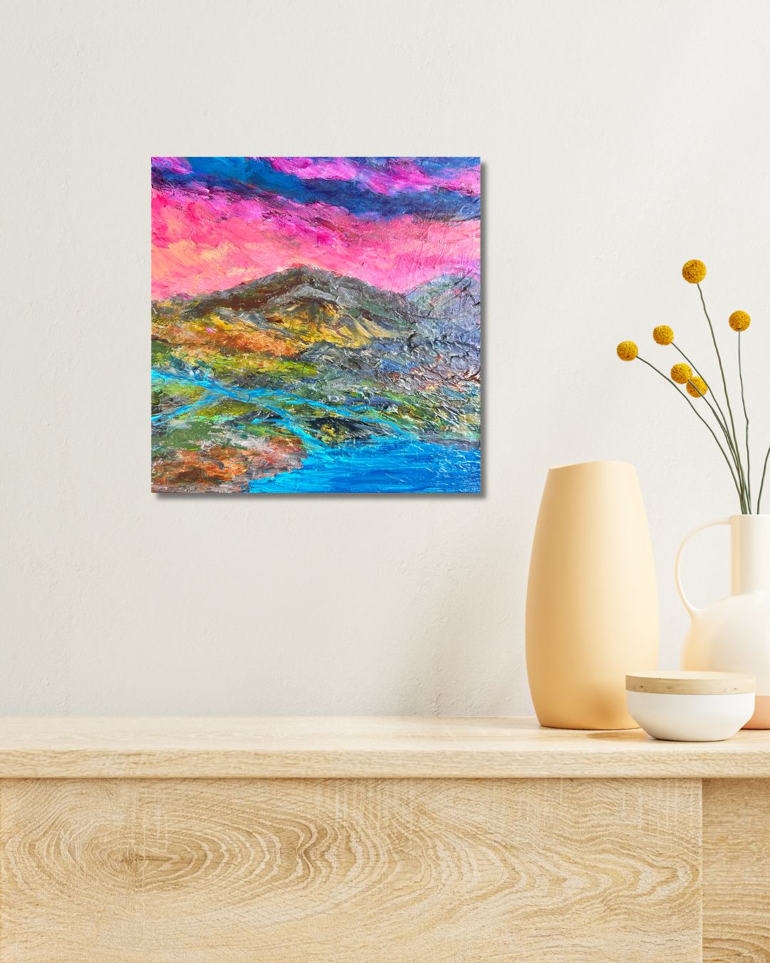 A colorful abstract landscape painting of mountains and a river under a vibrant pink and blue sky, by Ottawa artist Mireille Laroche, displayed on a light beige wall above a wooden console table with decorative vases.