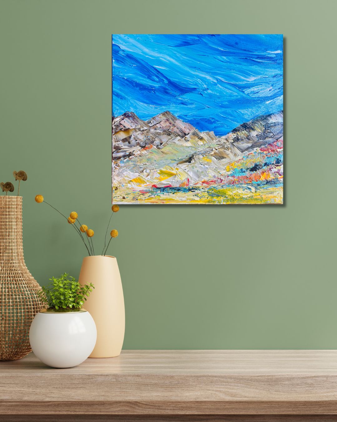 A colorful abstract mountain landscape painting with a bright blue sky by Ottawa artist Mireille Laroche displayed on a sage green wall above a wooden table with decorative vases and a small plant.