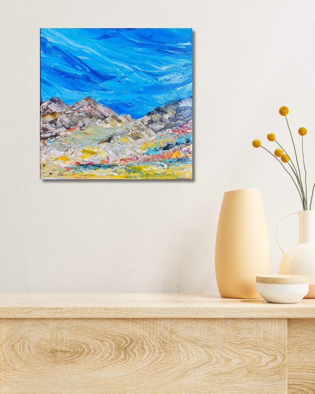 A colorful abstract mountain landscape painting with a bright blue sky by Ottawa artist Mireille Laroche on a creme wall above a dresser with vases on it