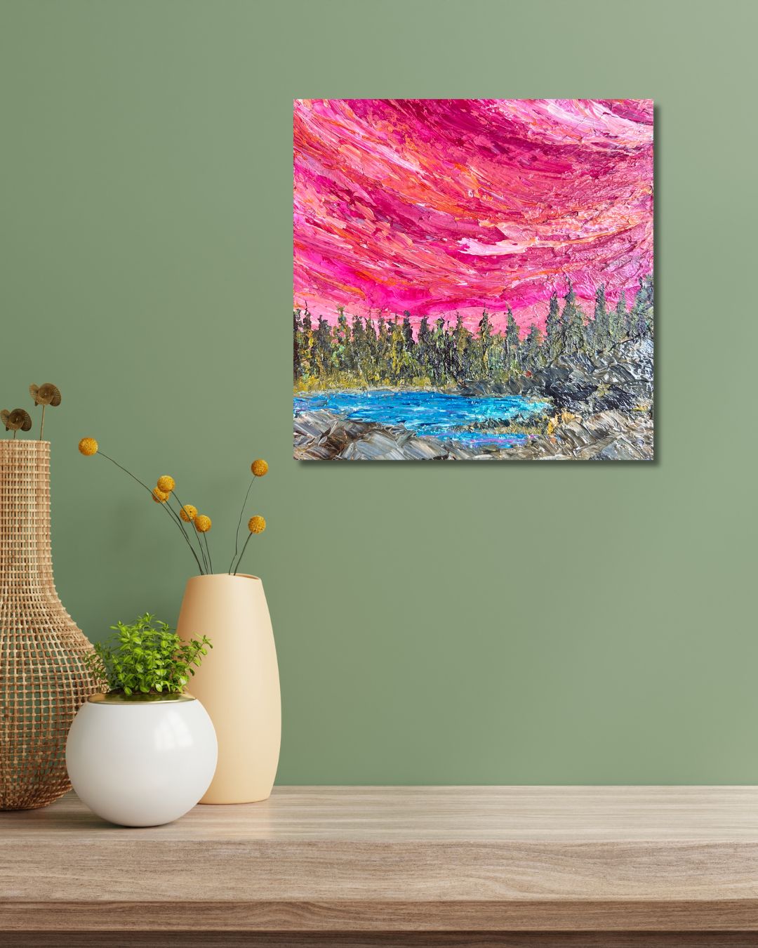 A square canvas painting of a vibrant pink and red sky over a blue lake and pine forest, by Ottawa artist Mireille Laroche, displayed on a sage green wall above a wooden table with decorative vases.