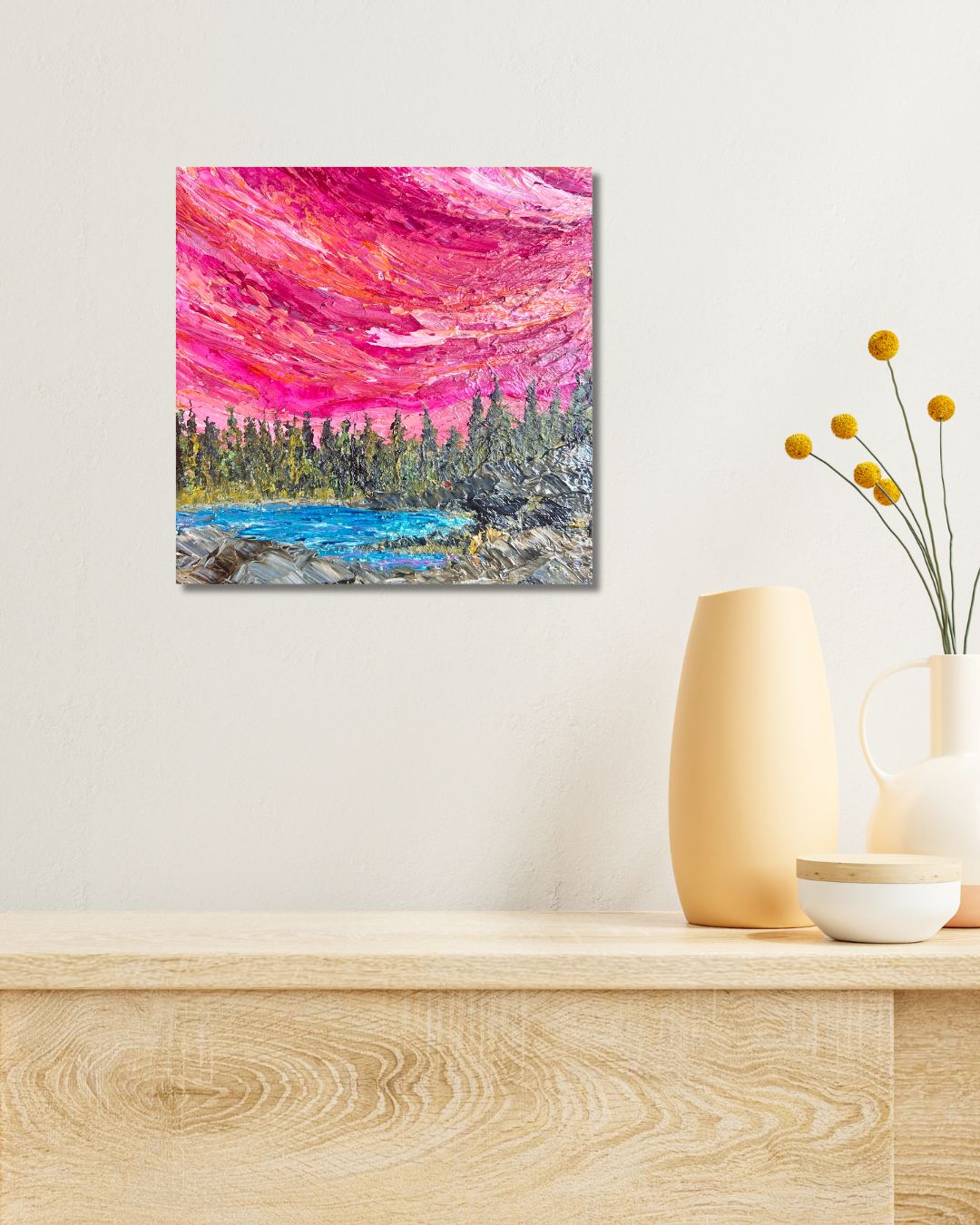 A square canvas painting depicting a vibrant pink and red sky over a blue lake and green forest, by Ottawa artist Mireille Laroche, displayed on a light beige wall above a wooden console table with decorative vases.