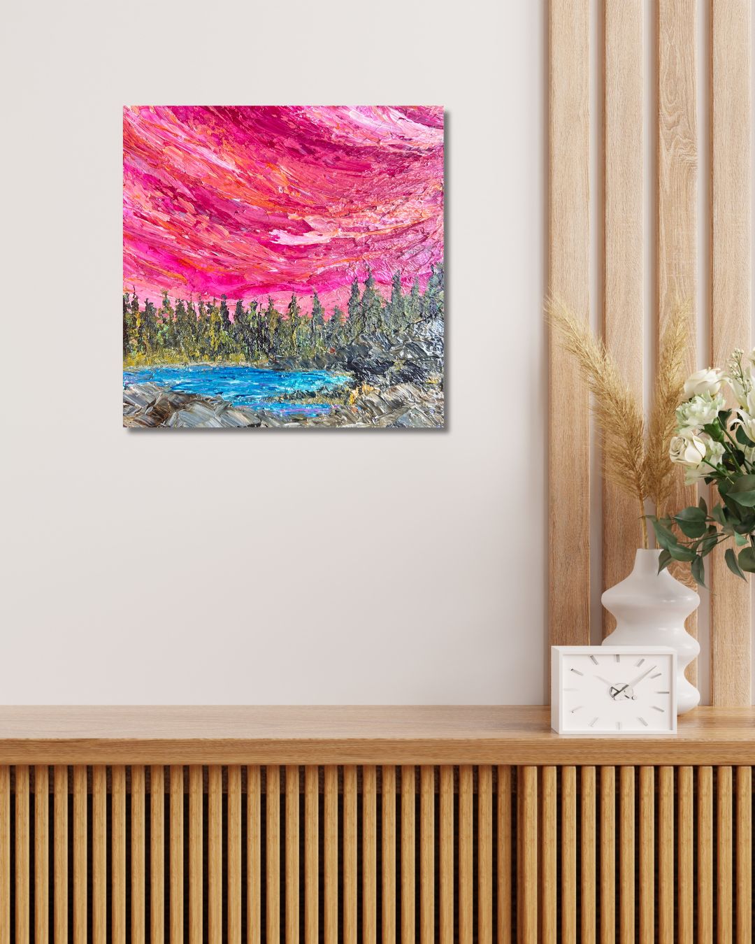 A square canvas  painting depicting a vibrant pink and red sky over a blue lake and green forest, by Ottawa artist Mireille Laroche, displayed on a light beige wall above a wooden console table with a white clock and vase.