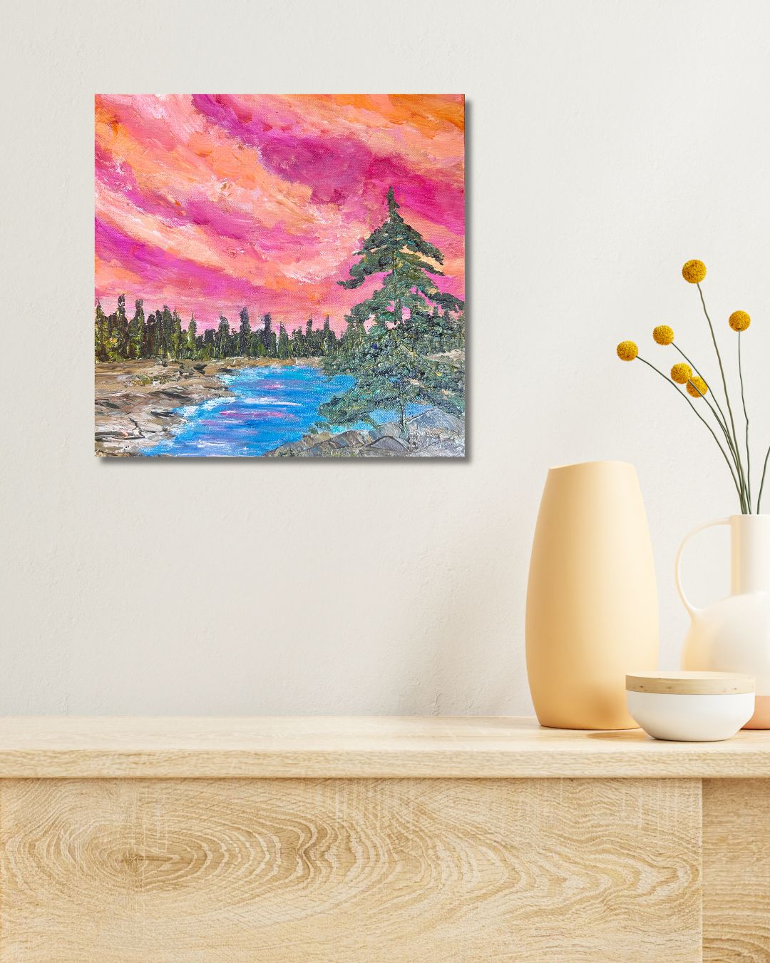 A square canvas artwork depicting a vibrant landscape with a pink and orange sunset sky, a blue river, and green pine trees, by Ottawa artist Mireille Laroche, displayed on a light beige wall above a wooden console table with decorative vases.
