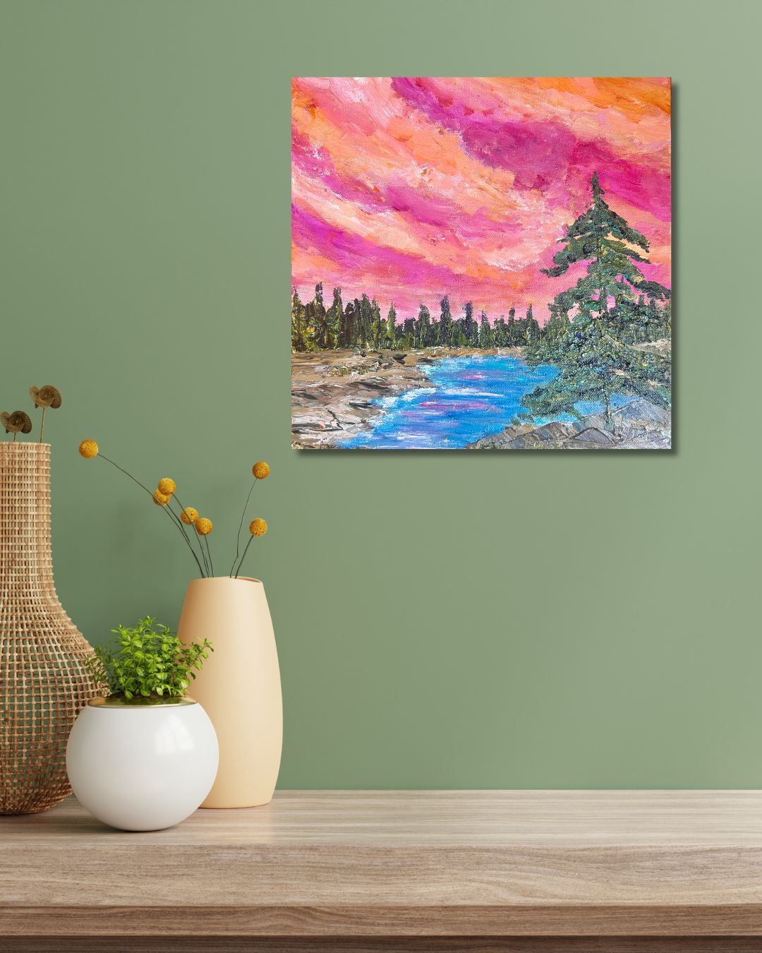 A colorful landscape painting of a river and pine trees under a vibrant pink and orange sky, by Ottawa artist Mireille Laroche, displayed on a sage green wall above a wooden table with decorative vases.
