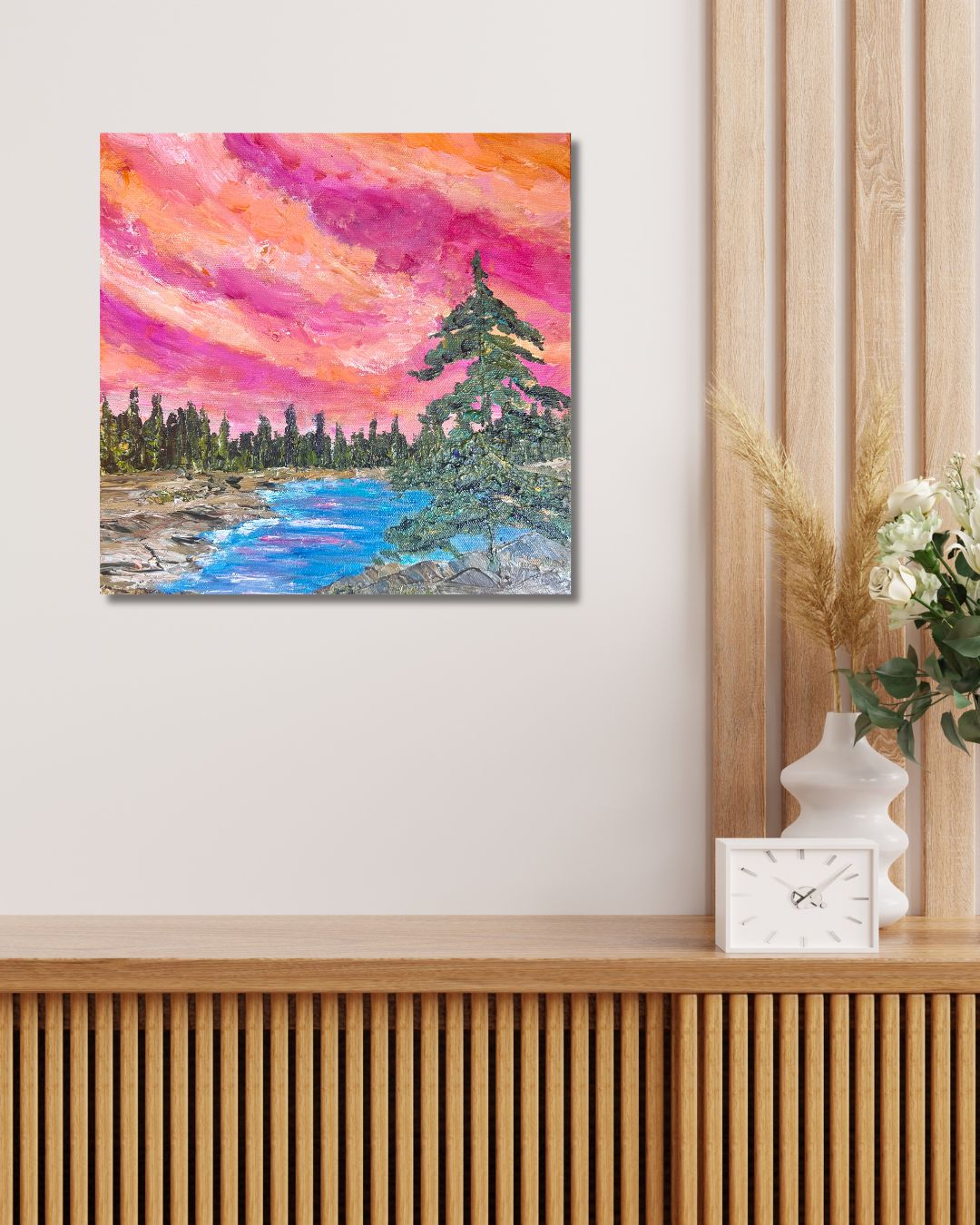 A square canvas painting of a colorful landscape with a pink and orange sky, blue river, and green trees, by Ottawa artist Mireille Laroche, displayed on a light wall above a wooden console table with a clock and vase.